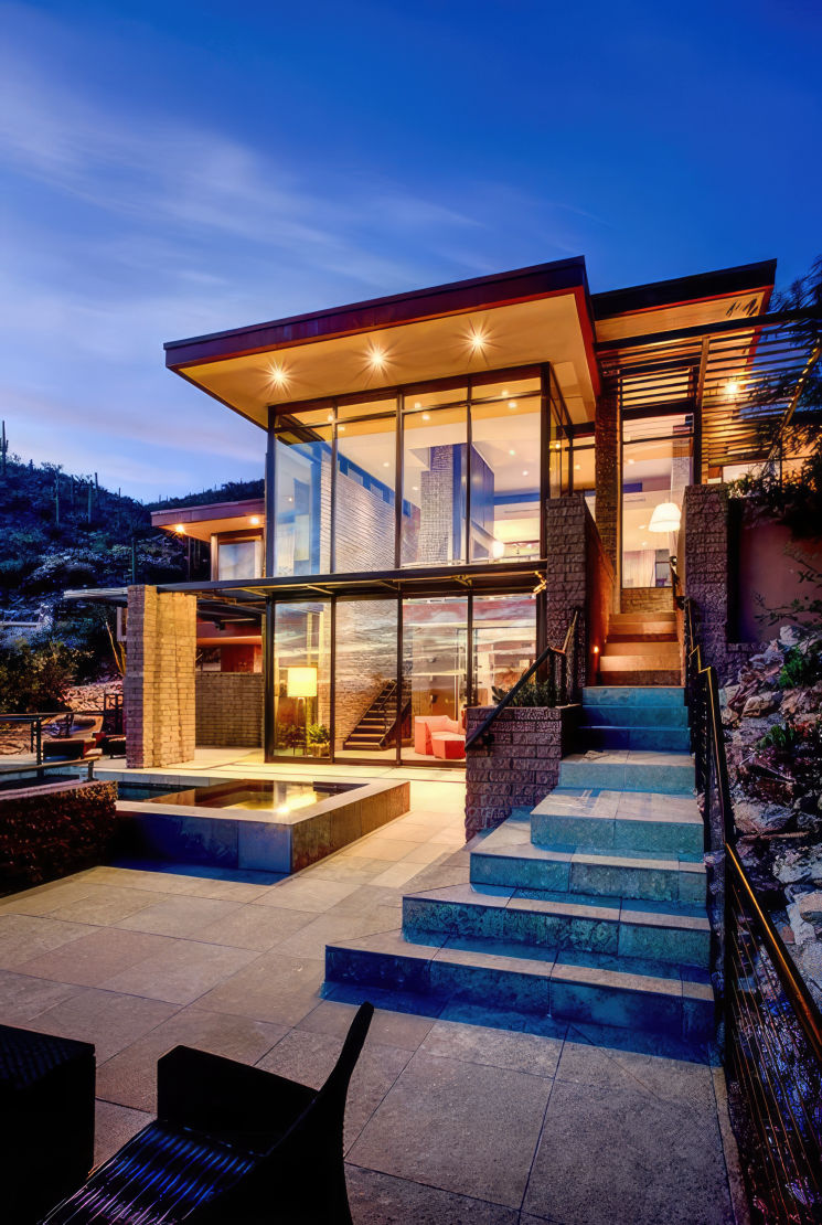 Fachada de una casa moderna con grandes ventanales y escalera exterior, iluminada al anochecer