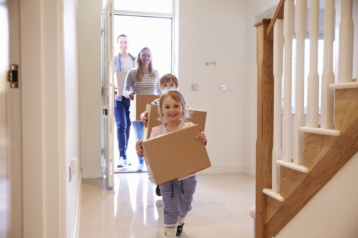 Familia entrando feliz en su nueva casa con cajas de mudanza, niños al frente y padres al fondo