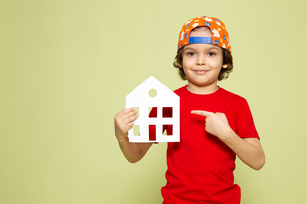 Niño sonriente con gorra hacia atrás señalando una casa de papel, representando el sueño de tener un hogar